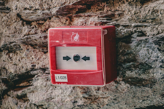 Close-up of a bright red manual fire alarm call point box mounted on a rough textured stone wall. The emergency push button features a flame icon and protective cover, ensuring safety and security