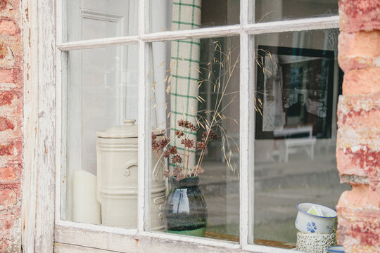 Horizontal view through an old paned window with peeling white paint, showing a rustic still life of a ceramic crock, dried flowers in a vase, and vintage items, evoking a cozy and nostalgic feeling