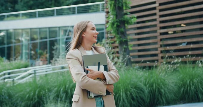 Beautiful young woman with natural smile, dressed in business casual attire, walks through sunny city park during her lunch break, looking directly at the camera with friendly and confident expression