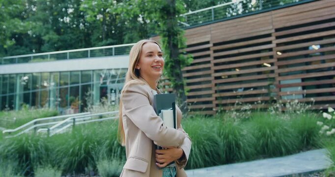 Side view of a young businesswoman with long hair in formal attire walking along the street near a modern office building while holding laptop, remote work, outdoors