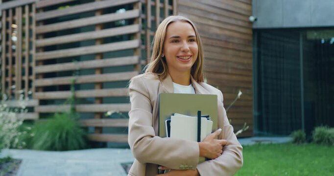 Young, beautiful, smiling woman with long hair walking outdoors in the city, looking at the camera, with an urban street in the background. Concept of confidence, freedom, and success.