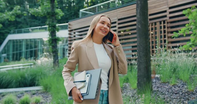 Confident businesswoman in a formal suit holding a laptop and talking on the phone while walking outdoors. Modern glass office building in the background. Business communication, important call