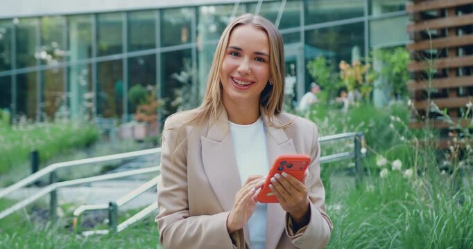 Close-up of portrait young woman using smartphone standing outdoors, looking at the camera with professional and approachable expression, dressed in casual clothes