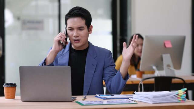 Stressed Asian businessman arguing on phone call at office desk. Manager is having an angry conversation on his mobile phone while sitting at his desk with a pile of paperwork in front of him.