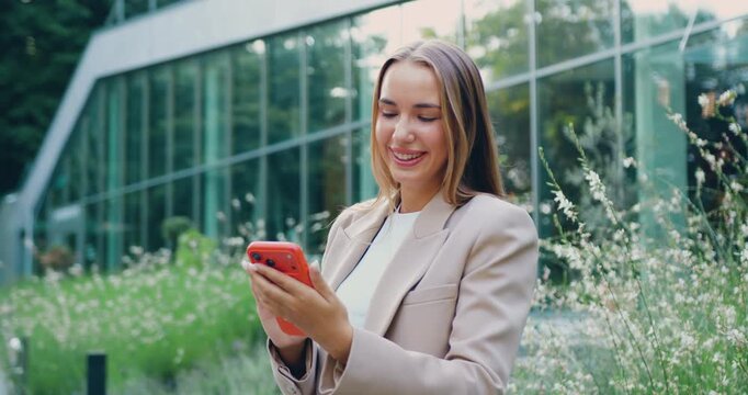 Young woman in formal business attire holding a mobile phone and looking to the side against the background of a modern glass office building.