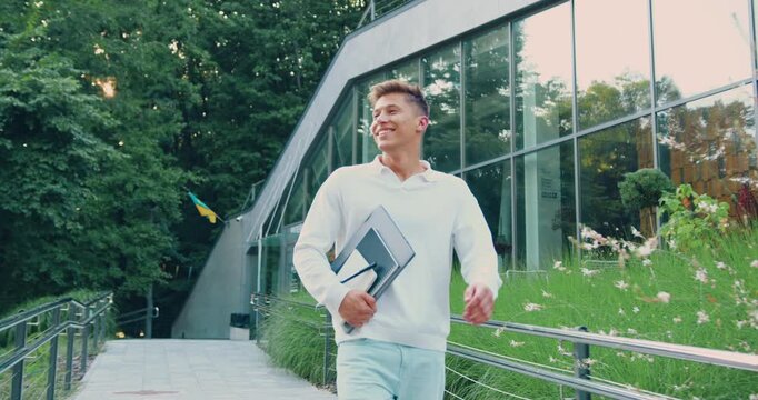 Side view of a young businessman in formal attire walking along the street near a modern office building while holding laptop, remote work, outdoors