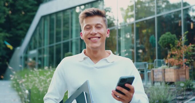 Portrait of smiling man employee in white shirt with smartphone walking outdoor. Young entrepreneur looking at camera along city street. Business career