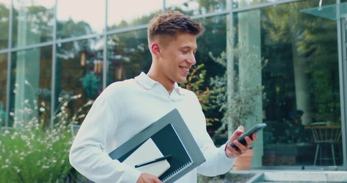 Man dressed in white shirt using smartphone using internet connection while walking outdoors on city street near a modern office building while holding laptop, remote work, outdoors