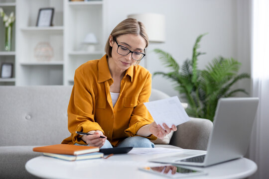 Woman wearing glasses and an orange shirt sitting on a sofa, holding papers and a pen, using a calculator and laptop to manage her personal finances and household budget