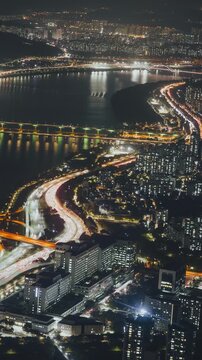 Aerial perspective of a vibrant city at night with a winding river, busy roads, and illuminated high-rise buildings visible. Time-lapse, Seoul, South Korea