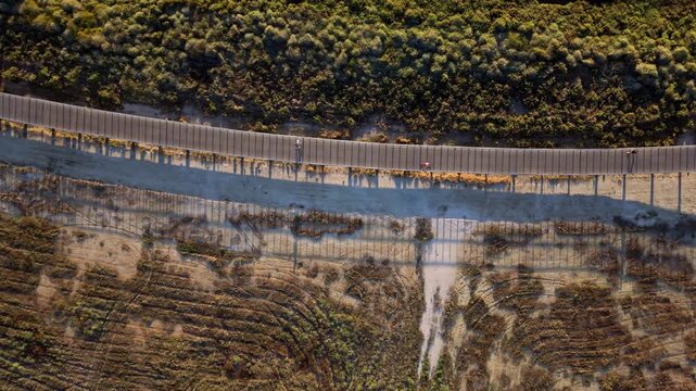 Aerial view shows long border fence stretching across hilly terrain. Fence runs parallel to road through dry, scrub-covered land. Sun casts long shadows over earthy slopes and vegetation