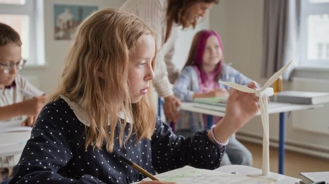 Caucasian child writing notes and spinning miniature wind turbine model during classroom lesson. Ideal for renewable energy education, sustainability projects and STEM school campaigns