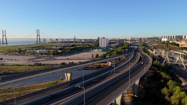 Aerial view of modern highway with curved roads. Vehicles move smoothly under clear blue sky. Bridge spans water in distance. City skyline visible behind greenery. Ideal for urban development