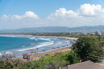 Pangandaran beach with golden sands curve along turquoise waves. White clouds drift above distant, hazy mountains. Beachgoers relax, swim, or stroll near boats. Blue sky contrasts with green foliage. © Rizky Rahmat Hidayat
