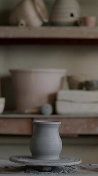 Traditional pottery workshop with clay vase on throwing wheel and handmade ceramics on wooden shelves. No people visible in background.