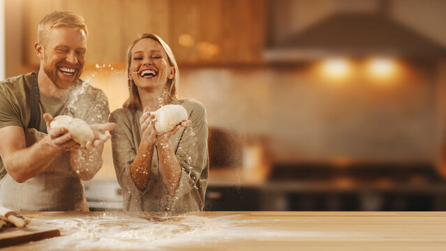 Laughing couple tossing pizza dough in flour with blurred warm kitchen background banner.