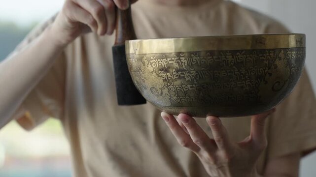Close up of a woman striking a Tibetan singing bowl and running a mallet along its rim to sustain tones. The etched surface and hand motion are in focus while the background is softly blurred Natural 