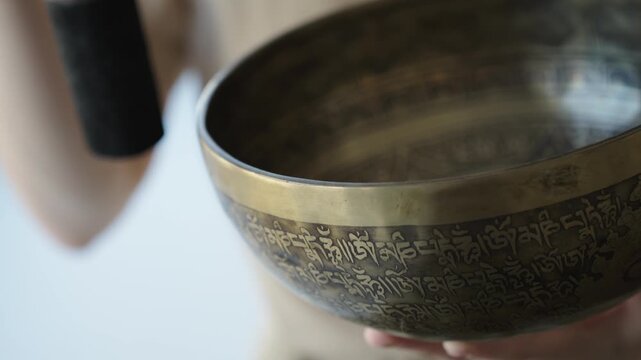 Close up of a woman striking a Tibetan singing bowl and running a mallet along its rim to sustain tones. The etched surface and hand motion are in focus while the background is softly blurred Natural 