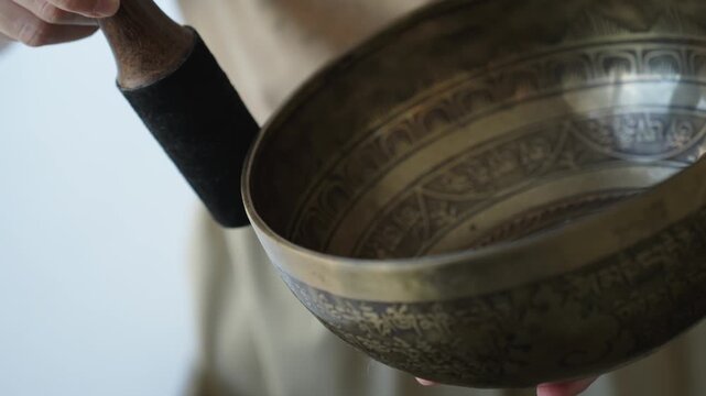 Close up of a woman striking a Tibetan singing bowl and running a mallet along its rim to sustain tones. The etched surface and hand motion are in focus while the background is softly blurred Natural 