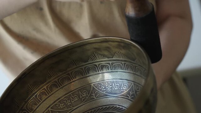 Close up of a woman striking a Tibetan singing bowl and running a mallet along its rim to sustain tones. The etched surface and hand motion are in focus while the background is softly blurred Natural 