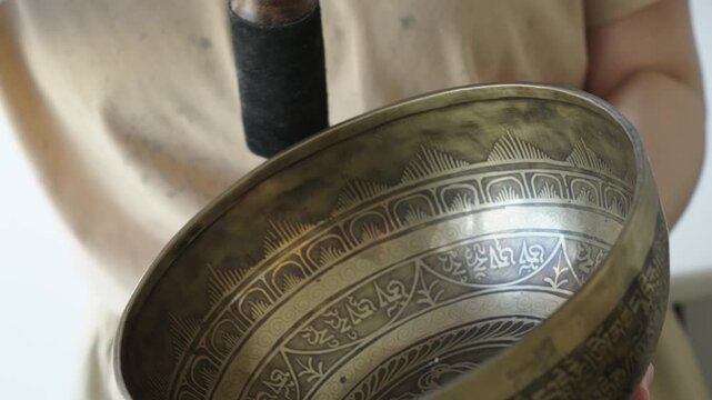 Close up of a woman striking a Tibetan singing bowl and running a mallet along its rim to sustain tones. The etched surface and hand motion are in focus while the background is softly blurred Natural 