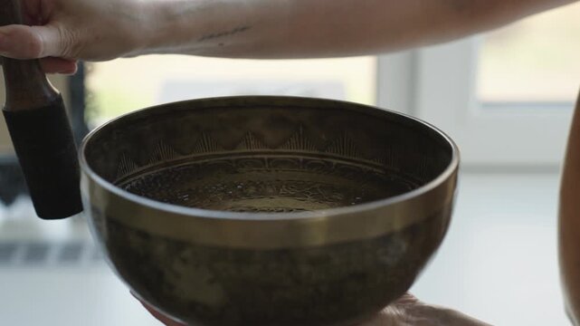 Close up of a hand striking a Tibetan singing bowl filled with water and running a mallet along the rim. The impact and rim motion create visible ripples and vibrating patterns on the liquid surface. 