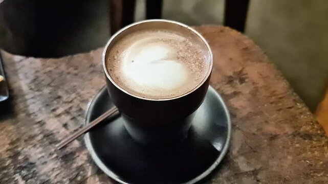 Hot cappuccino with heart latte art in a black ceramic cup and saucer on a rustic textured wooden table