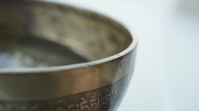 Close up of a hand striking a Tibetan singing bowl filled with water and running a mallet along the rim. The impact and rim motion create visible ripples and vibrating patterns on the liquid surface. 