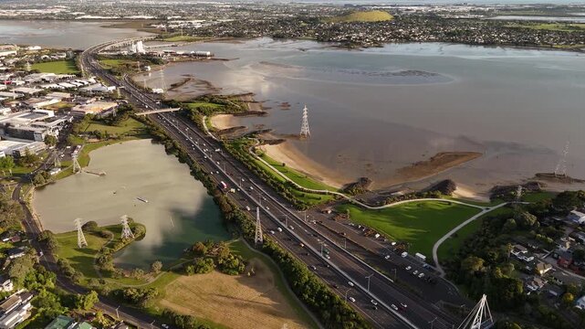 Multilane motorway curves across river estuary near Auckland suburb, cars stream through Mount Roskill interchange, muddy tidal flats stretch toward distant coastal hills. Aerial drone wide shot
