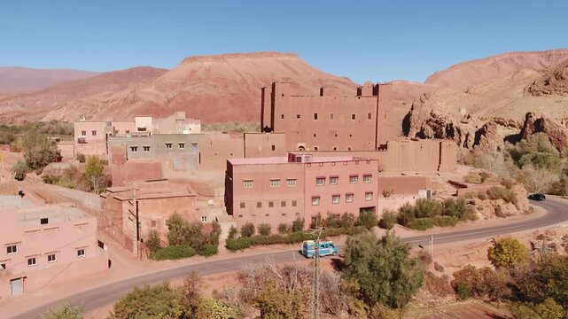 Elevated aerial shot following a blue local passenger minibus driving along a winding asphalt road past a traditional village and red mudbrick buildings in Tinghir, Morocco.