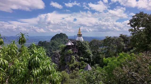 A panoramic timelapse video sweeping across the landscape of Wat Chaloem Phra Kiat, featuring a traditional white and gold pagoda perched on a rocky, forest-covered mountain peak under a cloudy blue s