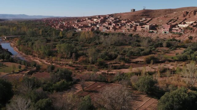 Scenic aerial panorama capturing the flowing Todgha river surrounded by dense green trees and a traditional mudbrick village on an arid hillside in Tinghir, Morocco.