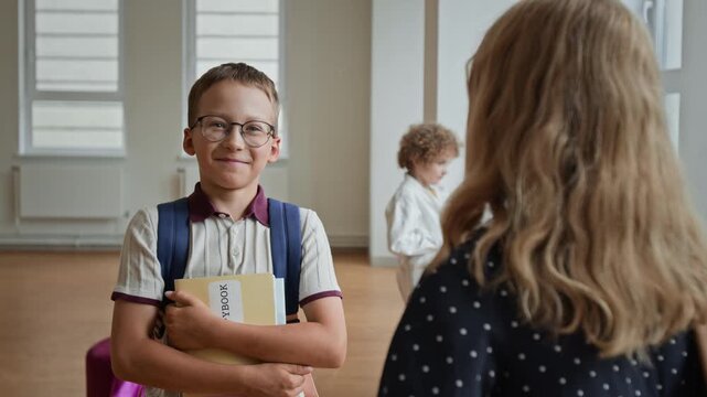 Caucasian schoolboy wearing eyeglasses holding copybook talking to classmate in hallway. Ideal for elementary education marketing, tutoring services, and academic ads