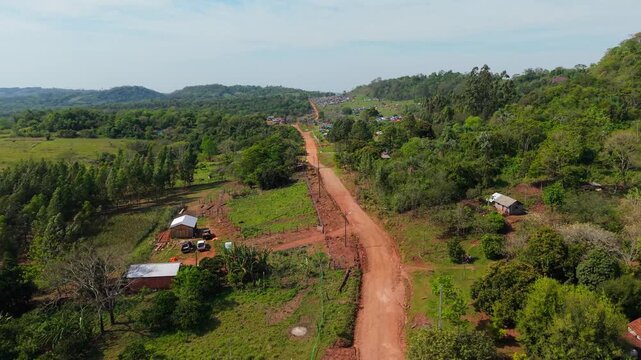 Drone shot flying along a winding red dirt road past rural farmhouses, leading toward a massive outdoor camping rally in Nueva Alborada, Paraguay