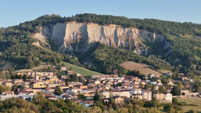 Monte Giogo rises above Lugagnano Val d'Arda with pale Pliocene clay cliffs, wooded slopes and cultivated fields in Piacenza, Emilia-Romagna, Italy, drone forward flyover