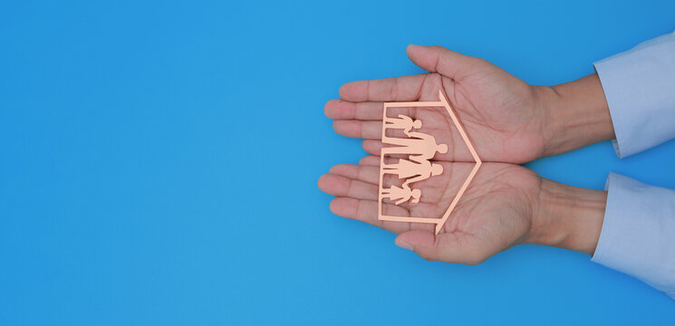 Caring hands hold wooden family cutout symbol for safe home ownership providing peaceful financial safeguard over blue background