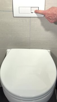 Vertical video social media - Closeup of a man&rsquo;s hand pressing a button on a tiled wall panel to flush the white bathroom toilet bowl below.