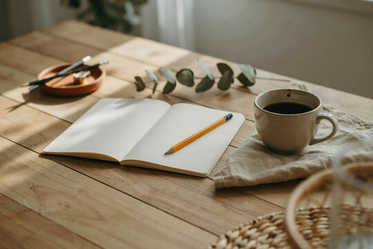 Open notebook with a yellow pencil on a wooden table, accompanied by a cup of black coffee, a small tray with pencils, and a green plant sprig