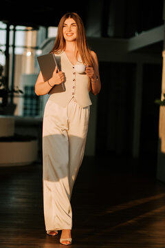Smiling businesswoman holding a laptop and cup while walking through a bright office during sunset