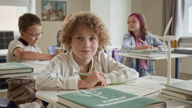 Portrait of Caucasian schoolboy sitting at desk with wind turbine model, smiling at camera in classroom. Footage suited for sustainability education campaigns and STEM learning promotions