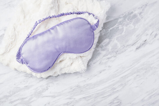 Silk purple sleep mask on soft fabric on white marble background, top view