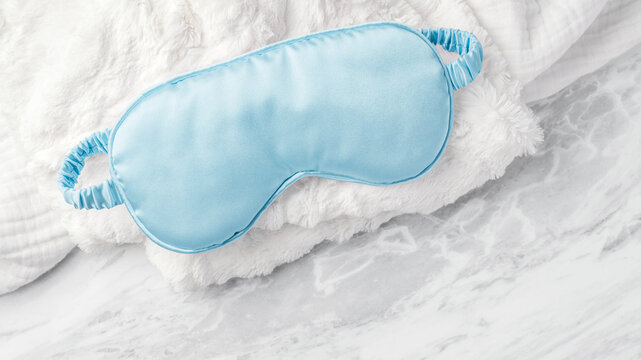 Blue sleep mask on soft fabric on white marble background, top view