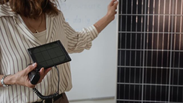 Caucasian middle aged female lecturer presenting compact solar panel beside large photovoltaic panel. Footage serves green energy education, sustainability advertising, and technology seminars