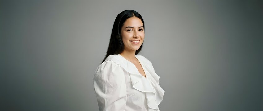 Young woman with long dark hair and South Asian ethnicity wearing a white ruffled blouse, smiling at camera against neutral gray background for corporate portraits and professional headshots