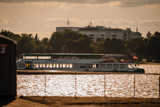 Passenger boat named "Ziemia" navigates a shimmering river with a backdrop of trees and a residential building under a cloudy sky during sunset