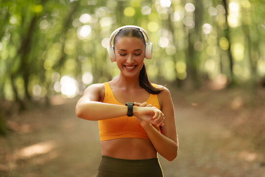 Young woman using wireless headphones and checking fitness smartwatch during outdoor training