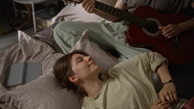 Young adult Caucasian woman resting in bed while listening to partner playing acoustic guitar, high angle view. Footage fits marketing campaigns on romance, leisure hobbies and mental wellbeing