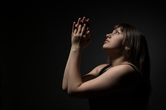 Young woman praying with hands clasped in profile view against a dark background