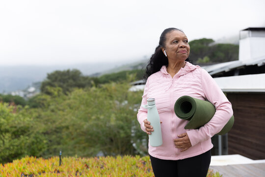 Senior African American woman in pink jacket standing on deck holding mat and bottle, copy space