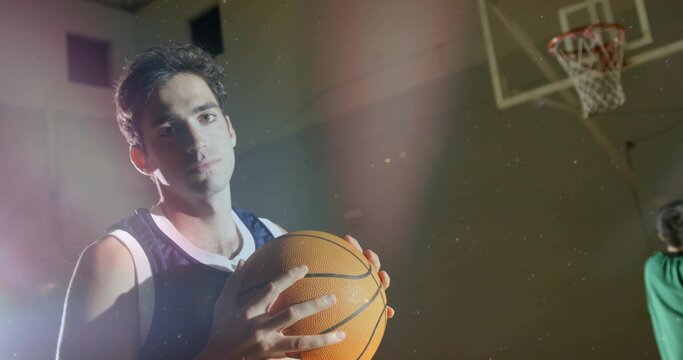 Holding basketball, male player standing in dim gym, wearing dark jersey, hoop visible